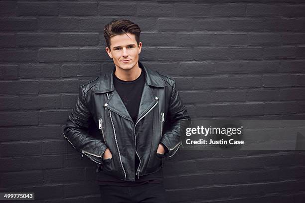 man in a leather jacket leaning on a brick wall - giacca di pelle foto e immagini stock
