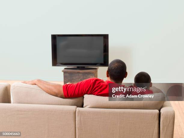 father and son watching television in living room - family watching tv from behind stockfoto's en -beelden