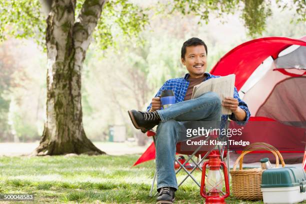 asian man relaxing in lawn chair at campsite - campingstuhl stock-fotos und bilder