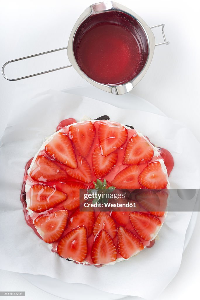 Pot of red cake glaze and strawberry cream cheese tart on white ground, elevated view