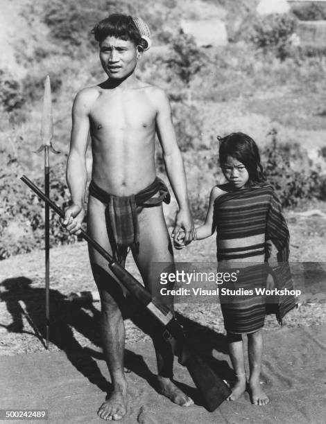 An Igorot tribe hunter poses with a young girl while displaying his gun and his spear, Luzon, Philippines, early to mid 20th century. The hunter...