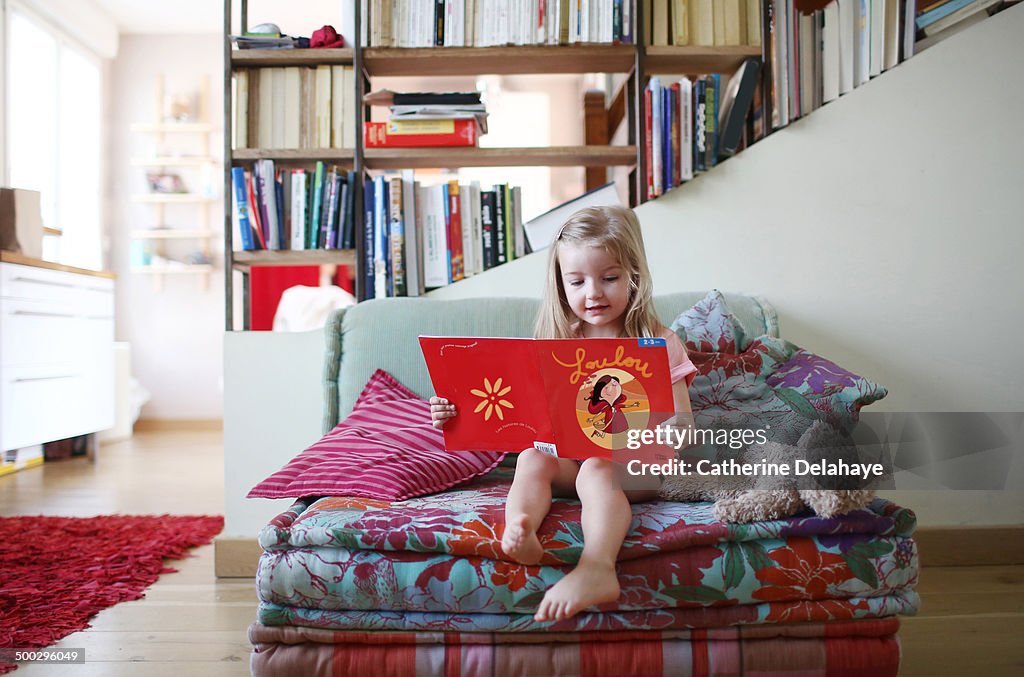 A 3 years old girl reading a book on a sofa