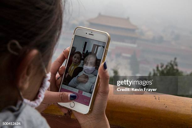 Chinese tourist looks at her phone after taking a picture of herself in a mask while visiting Jingshan Park overlooking the Forbidden City in heavy...