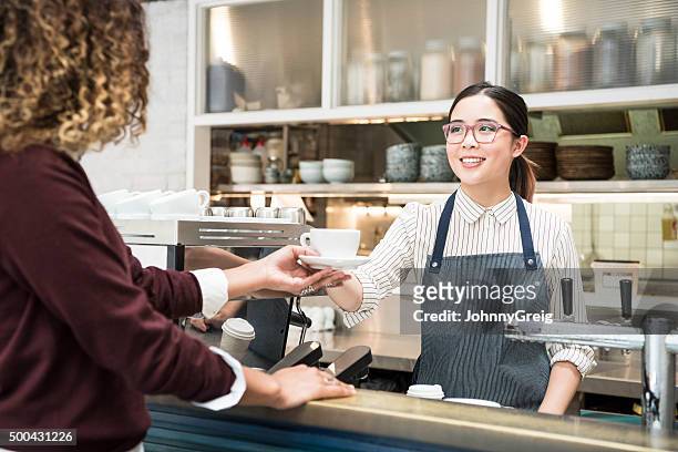 young asian barista serving coffee to female customer in cafe - coffee shop owner stockfoto's en -beelden