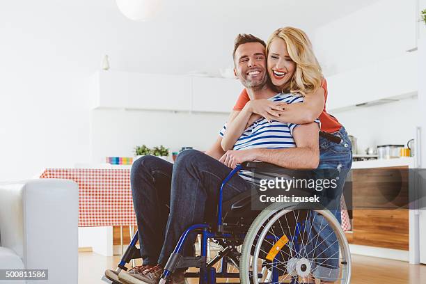behinderten mann sitzt im rollstuhl, seine frau umarmt ihn - rollstuhl zu hause stock-fotos und bilder