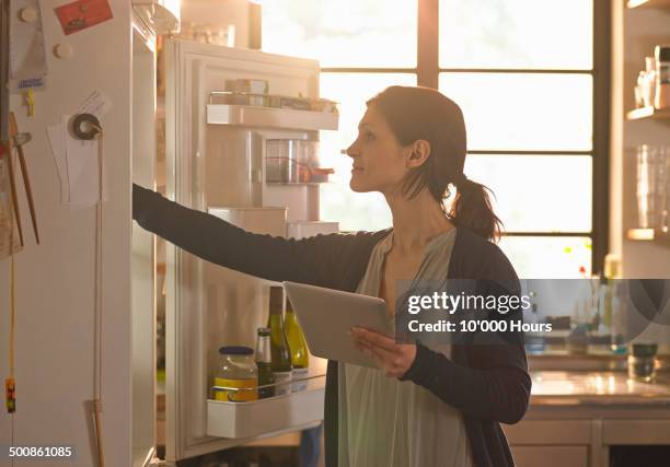 woman holding an tablet computer whilst looking in the fridge - protocolo fotografías e imágenes de stock