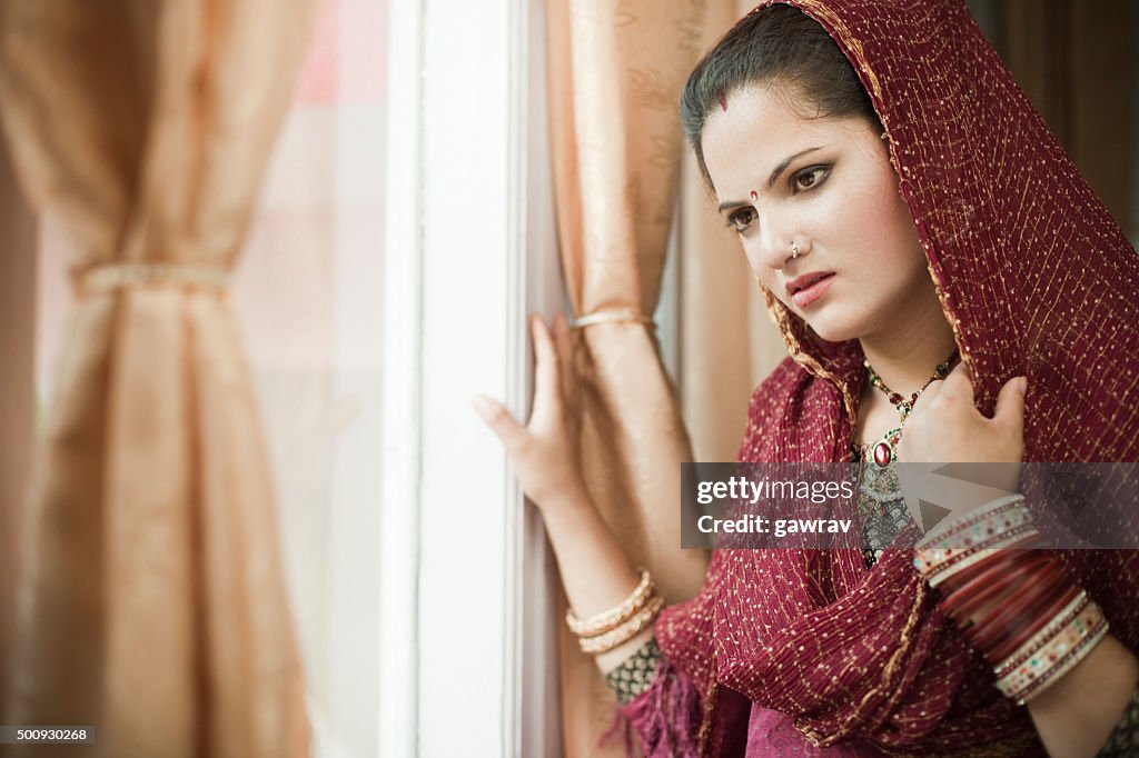 Beautiful newly married sad Indian young woman standing near window.