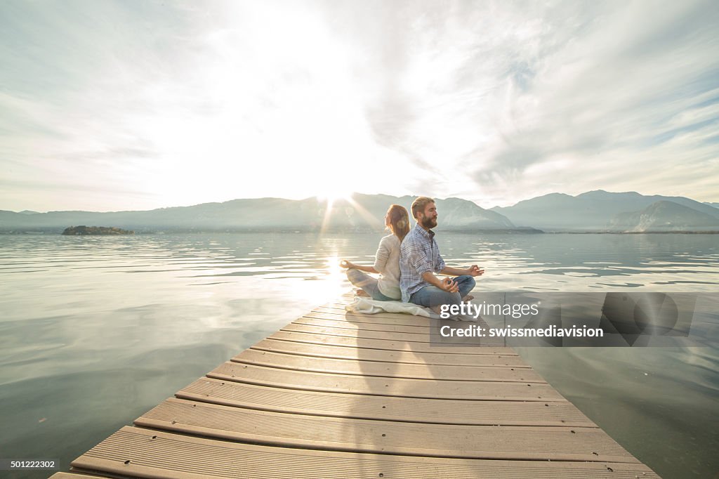 Young couple sits on jetty above lake, exercises yoga