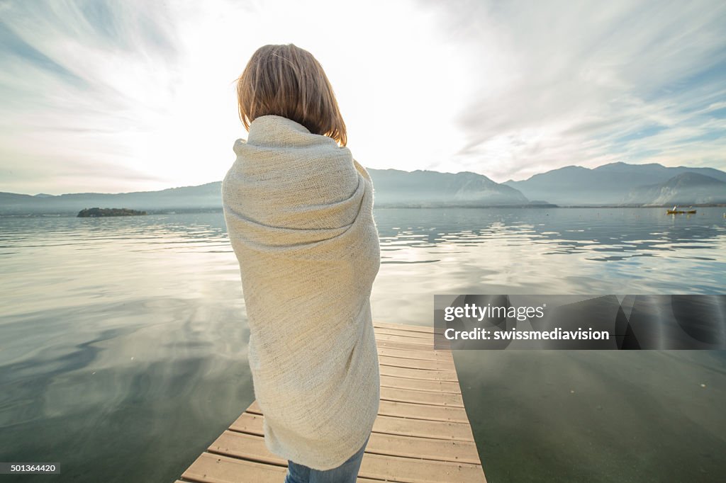 Woman watches sunset from lake pier, wrapped in blanket