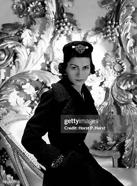 Coco Chanel wearing one of her designs: a three-quarter coat with cinched waist and astrakhan lapels and cuffs, and matching astrakhan hat with large...
