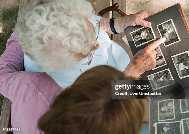 senior woman sitting on park bench with granddaughter, looking at old photograph album - album de fotos fotografías e imágenes de stock