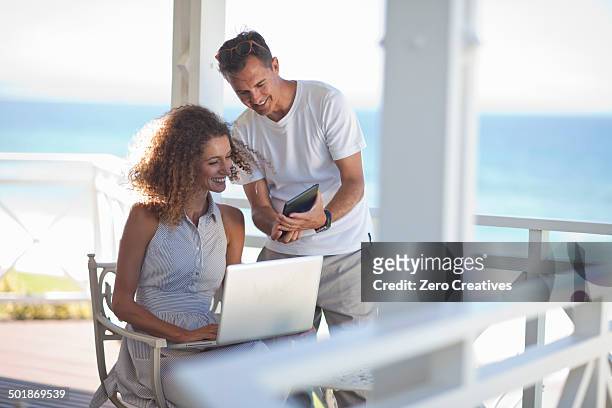 couple looking at digital tablet and laptop on beach house balcony - beach house balcony stock pictures, royalty-free photos & images