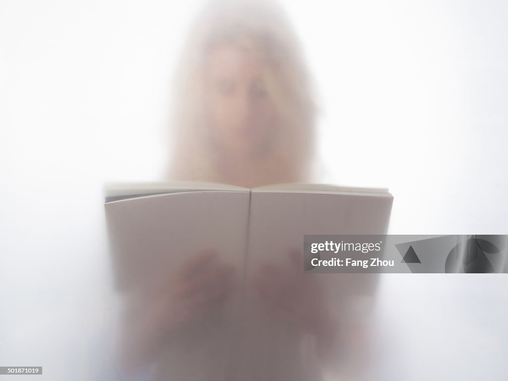 Young woman reading book behind frosted glass
