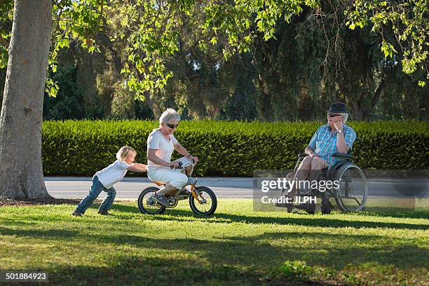 three year old boy pushing grandmother on cycle with grandfather watching from wheelchair - 70 year male stock pictures, royalty-free photos & images