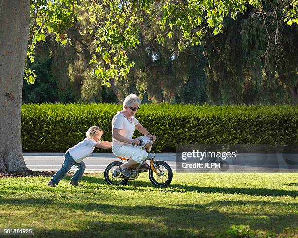 three year old boy pushing grandmother on cycle in park - velo humour stock-fotos und bilder