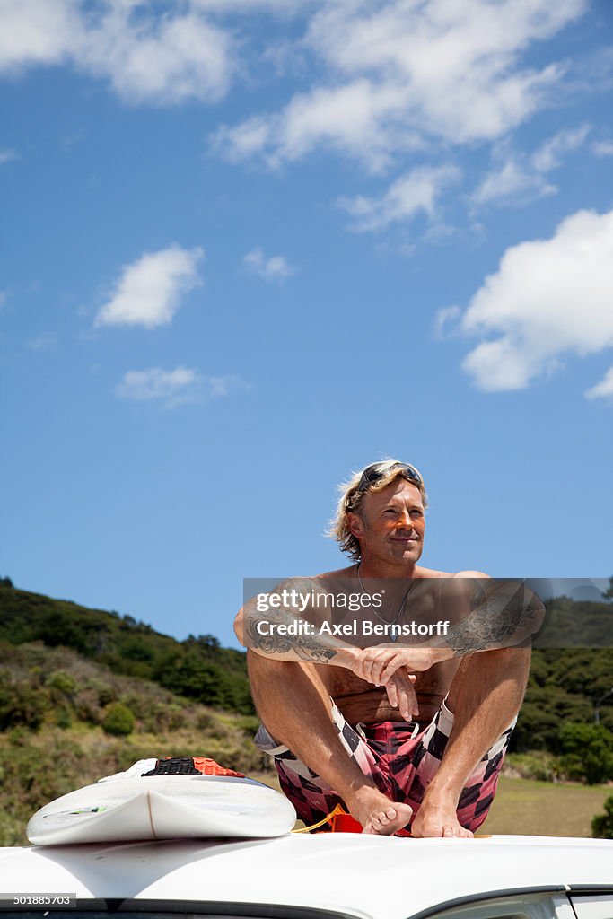 Mature man with surfboard sitting on vehicle roof at beach