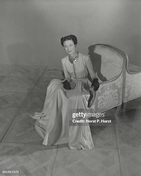 The Duchess of Windsor, sitting in an armchair and wearing a cap-sleeved, silk shantung gown by Mainbocher with black satin gloves and a gold...