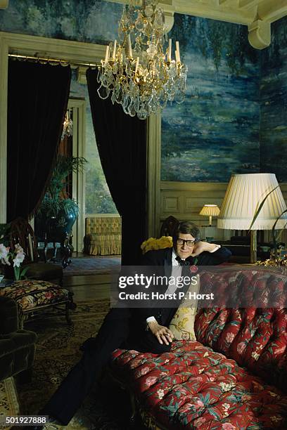 Fashion designer Yves Saint Laurent wearing dark eyeglasses and a tuxedo, holding a cigarette in his right hand while reclining on a floral settee at...