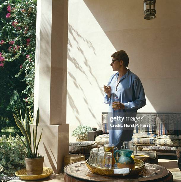 Designer Yves Saint Laurent, in profile, wearing a blue caftan standing in his Marrakesh patio holding a glass and a cigarette.