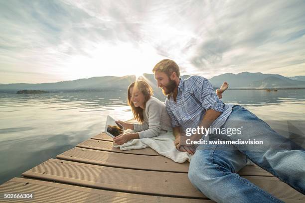 young couple lies on jetty above lake, uses digital tablet - couple jetty stock pictures, royalty-free photos & images