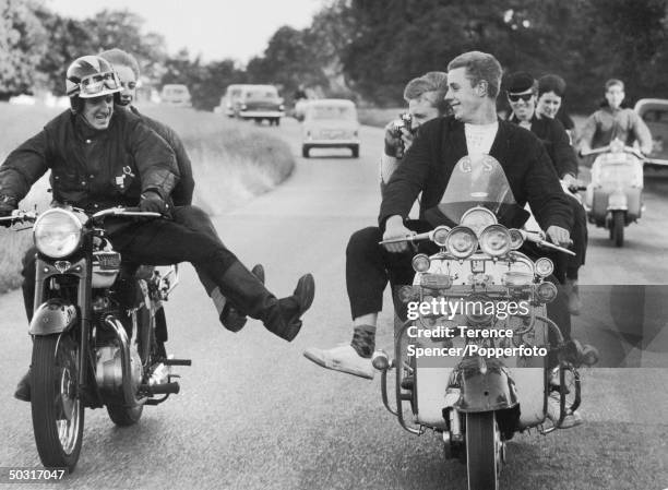 Pair of Rockers on a Triumph motorbike joke and jest with a convoy of mods riding scooters along a road running through a park in England in July...