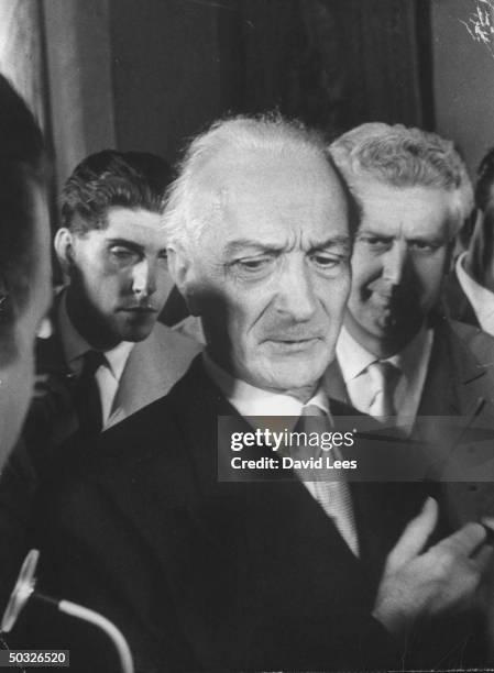 Italian Prime Minister Antonio Segni standing at the microphone while being surrounded by newsmen interviewing him.