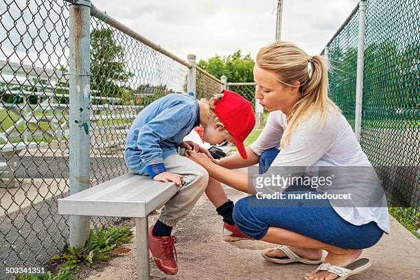 mom and son blowing on scraped knee on baseball bench. - ehbo stockfoto's en -beelden