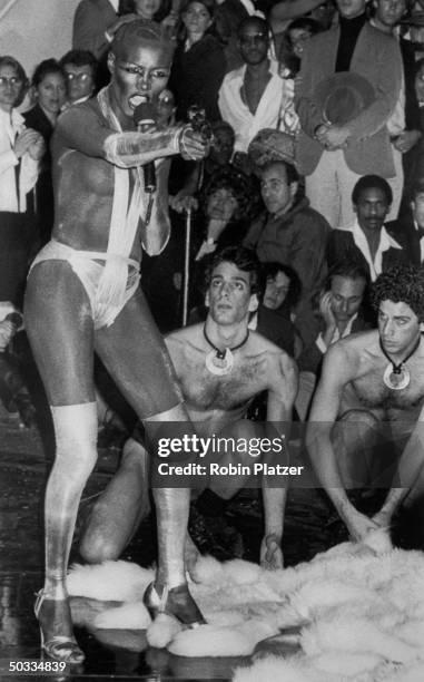 Singer Grace Jones wearing a see-through body stocking and not much else performing a song w. Near-naked men at her side at Studio 54.