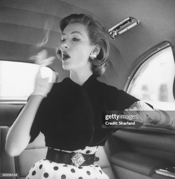 Model wearing velvet bolero jacket over polka-dotted sheath dress, smoking in back seat of automobile.