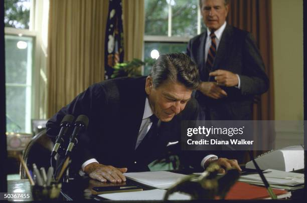 Vice Pres. George H. W. Bush watching as Pres. Ronald W. Reagan reads a note on his desk in the Oval Office.