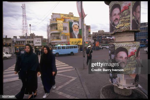 Women crossing street by pole plastered w. Presidential election campaign posters for moderate front-runner Mohammed Khatami, banner imaging...