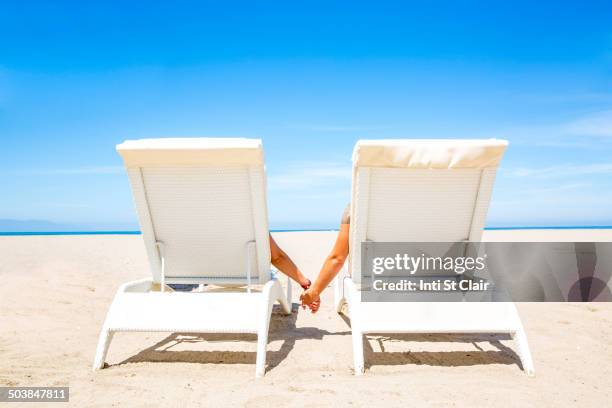 couple holding hands in lounge chairs on beach - partie-médiane photos et images de collection