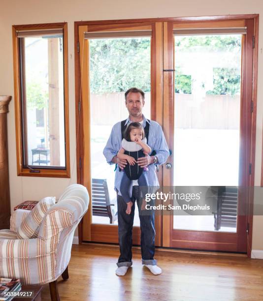 father holding daughter in sling in living room - baby carrier stock pictures, royalty-free photos & images