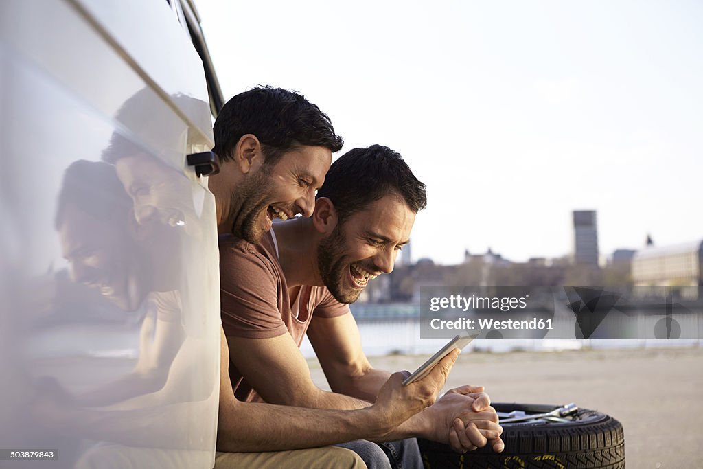 Two laughing men sitting in car looking at digital tablet