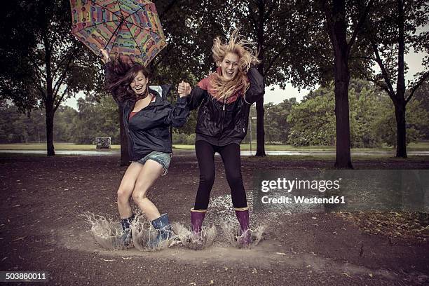 two playful young women with umbrella jumping in puddle - friends prank stock pictures, royalty-free photos & images