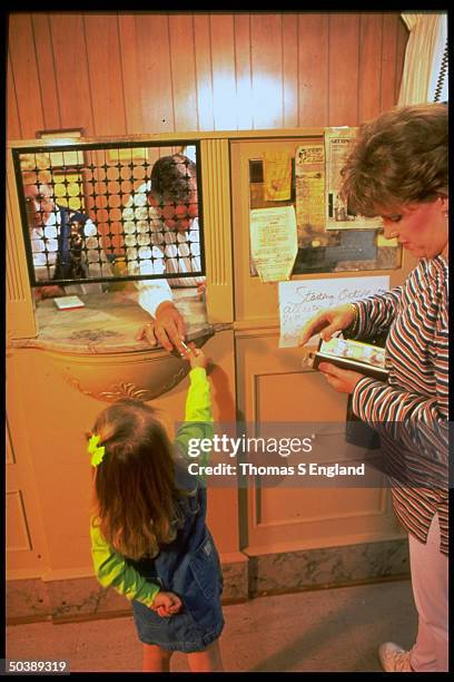 Bank teller Jack Wilkerson handing a lollipop through his window to Mercedes Smith as her mom Angie looks on , while banking at the Bank of Lowes,...