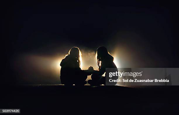 silhouette of two girls sitting on the pavement of a road, facing, holding hands in the dark - friends loneliness stock-fotos und bilder