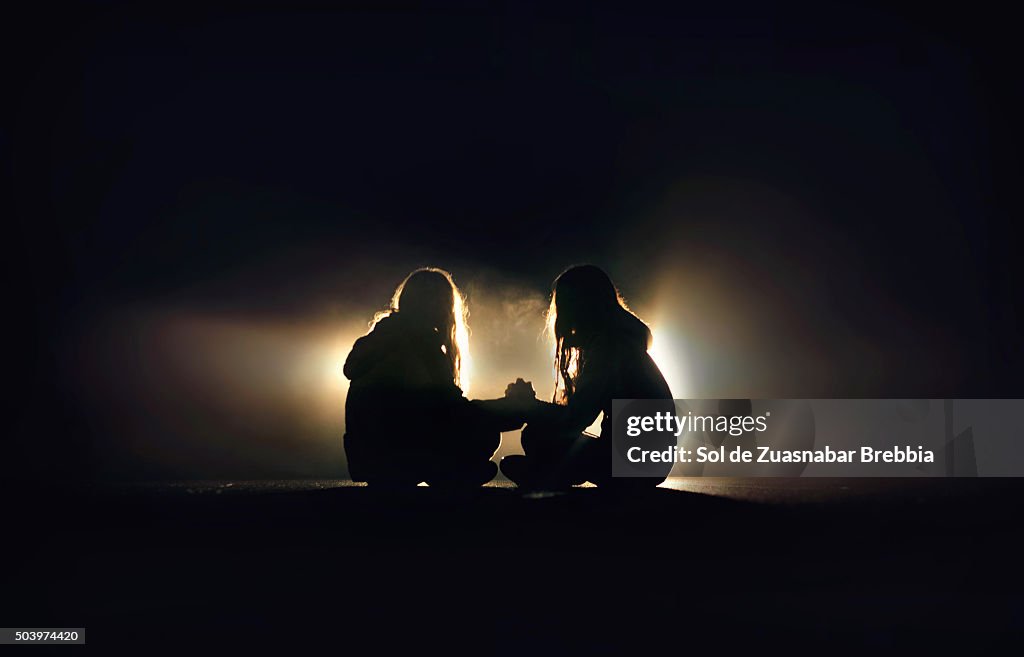 Silhouette of two girls sitting on the pavement of a road, facing, holding hands in the dark