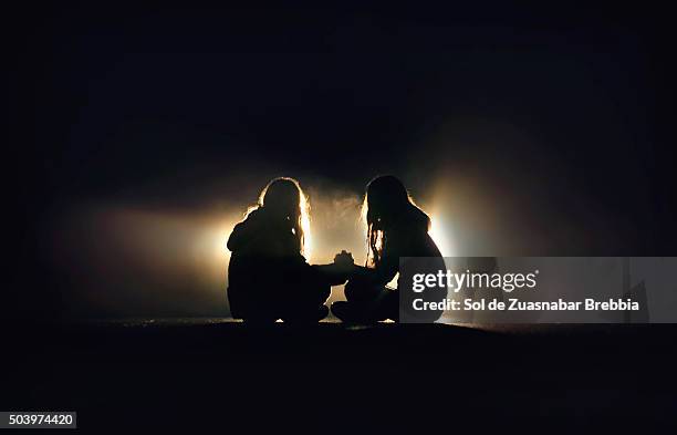 silhouette of two girls sitting on the pavement of a road, facing, holding hands in the dark - friends loneliness imagens e fotografias de stock