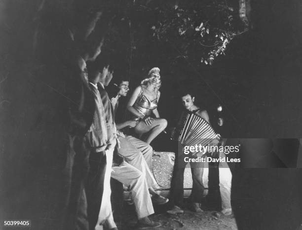 Singer Juli Reding standing in her car singing while a man plays the accordion for a group of fans during her visit.