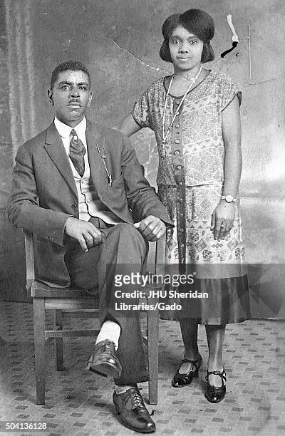 Young African American couple, a man seated in a chair and wearing a suit, woman standing and wearing a dress, watch, and patent leather shoes with...
