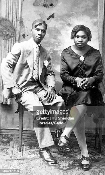 Young African American couple, a man and a woman seated together on a bench, the man wearing a suit, the woman wearing a dress and patent leather...