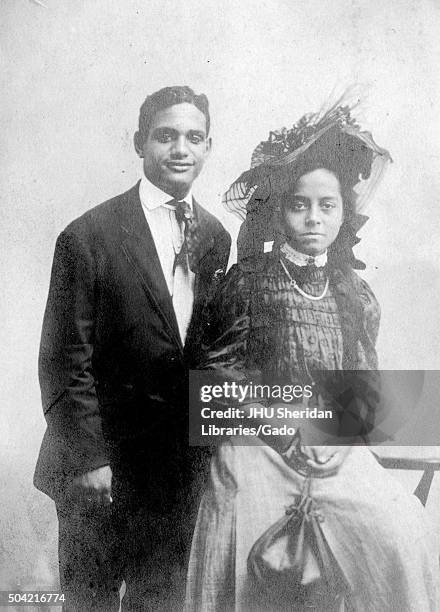 African-American couple posing for a photograph, the man wearing a suit and smiling, the women sitting on a chair in front of the man, wearing a hat,...