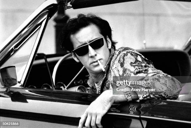 Actor Nicolas Cage, wearing sunglasses and snakeskin jacket, w. Cigarette hanging out of his mouth as he sits in convertible during filming of movie...