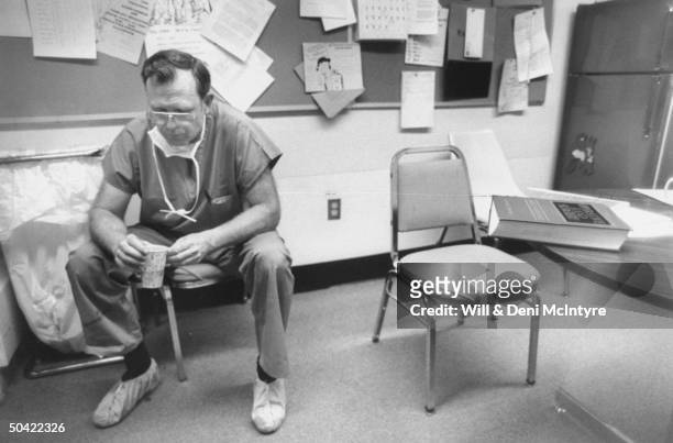 Hand Surgeon Dr. Abner Bevin in surgical scrubs slumped in chair w. Cup in hand, prob. After performing hand surgery on Carpal Tunnel Syndrome...