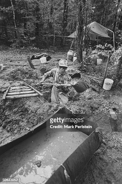 Diamond prospector Jim Archer digging deep hole in the mud w. Bucket shovel as he fills nearby trough w. Slop to be screened for diamonds at Crater...