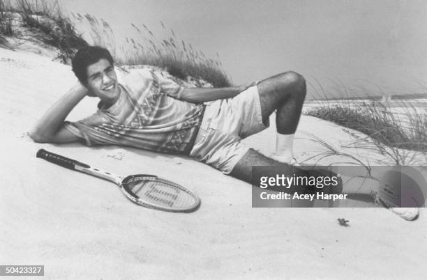 Open tennis champ Pete Sampras wearing t-shirt & shorts, stretched out on the sand next to a wooden Wilson tennis racket at isolated beach.
