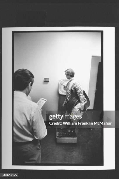 Jockey Pat Day wearing his racing silks as he stands on scales holding his saddle & gear during weigh-in as man looks on in jockey room before race...