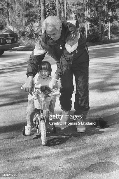 Sportscaster Pat Summerall pushing his 4-yr.-old granddaughter Caroline on her little tricycle w. Basket on the handlebars containing a darling puppy...