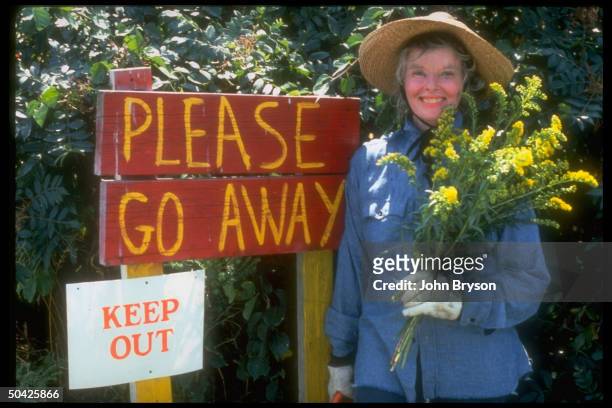 Actress Katharine Hepburn wearing jean shirt & sun-hat, holding flowers next to homemade sign saying Go Away in her driveway.